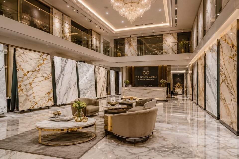 Ultra-luxury imported marble showroom interior, massive premium white marble slabs displayed elegantly, high-end marble gallery featuring Statuario Marble, Carrara Marble, Calacatta Gold Marble, Volakas Marble, Thassos White Marble, sophisticated Italian marble showroom in Kishangarh, modern luxury architecture, double-height ceiling, warm ambient lighting, polished marble flooring reflecting light, designer marble walls, premium luxury interiors, elegant reception counter, stylish lounge seating, rich golden accents, contemporary showroom design, cinematic lighting, ultra-realistic textures, luxury natural stone atmosphere, modern chandelier lighting, premium imported marble display racks, luxurious commercial interior, architectural visualization, hyper-realistic 8K render, elegant modern luxury showroom aesthetics, spacious premium marble experience center, refined interior styling, photorealistic luxury marble showroom scene